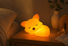 `Veilleuse en forme de renard endormi allumée diffusant une lumière chaude orange sur une table de chevet en bois`