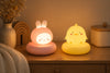 Veilleuses lumineuses en forme de lapin rose et de poussin jaune posées sur une table de chevet en bois