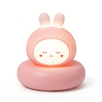 Veilleuse en forme de tête de petit lapin rose allumée, posée sur un large socle circulaire assorti sur fond blanc uni. Le personnage présente un visage blanc lumineux et arrondi avec des yeux clos, un petit nez rouge et deux petits cœurs dessinés sur les joues, tandis que ses deux petites oreilles dressées diffusent également une douce lueur chaude.