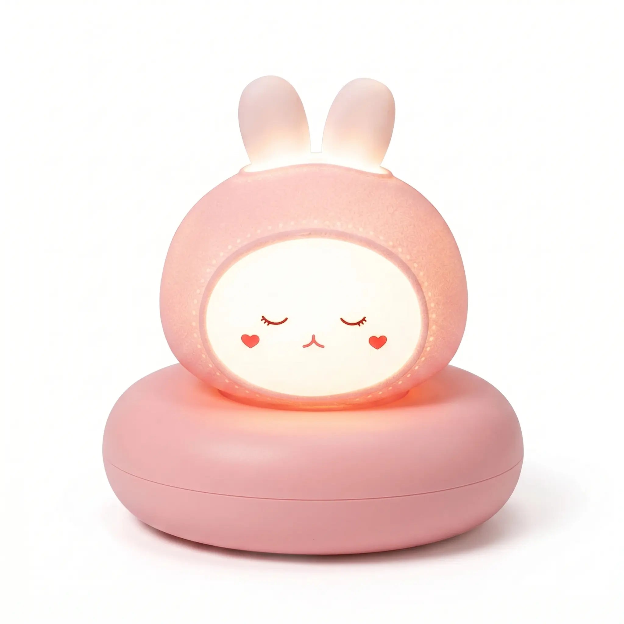 Veilleuse en forme de tête de petit lapin rose allumée, posée sur un large socle circulaire assorti sur fond blanc uni. Le personnage présente un visage blanc lumineux et arrondi avec des yeux clos, un petit nez rouge et deux petits cœurs dessinés sur les joues, tandis que ses deux petites oreilles dressées diffusent également une douce lueur chaude.