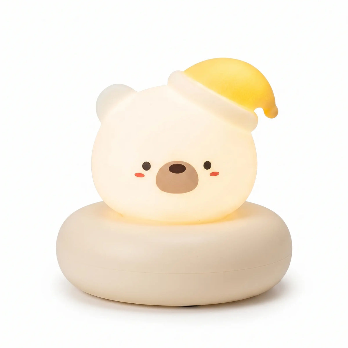 Veilleuse en forme de tête de petit ours blanc allumée, posée sur un large socle circulaire assorti de couleur crème sur fond blanc uni. Le personnage présente un visage blanc lumineux et arrondi avec des petits yeux noirs, un museau marron, des joues rosées et porte un bonnet de nuit jaune incliné sur le côté, diffusant une douce lueur chaleureuse.