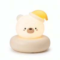 Veilleuse en forme de tête de petit ours blanc allumée, posée sur un large socle circulaire assorti de couleur crème sur fond blanc uni. Le personnage présente un visage blanc lumineux et arrondi avec des petits yeux noirs, un museau marron, des joues rosées et porte un bonnet de nuit jaune incliné sur le côté, diffusant une douce lueur chaleureuse.