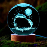 Veilleuse boule de cristal dauphin éclairée en bleu posée sur un socle en bois au cœur d'un décor naturel verdoyant