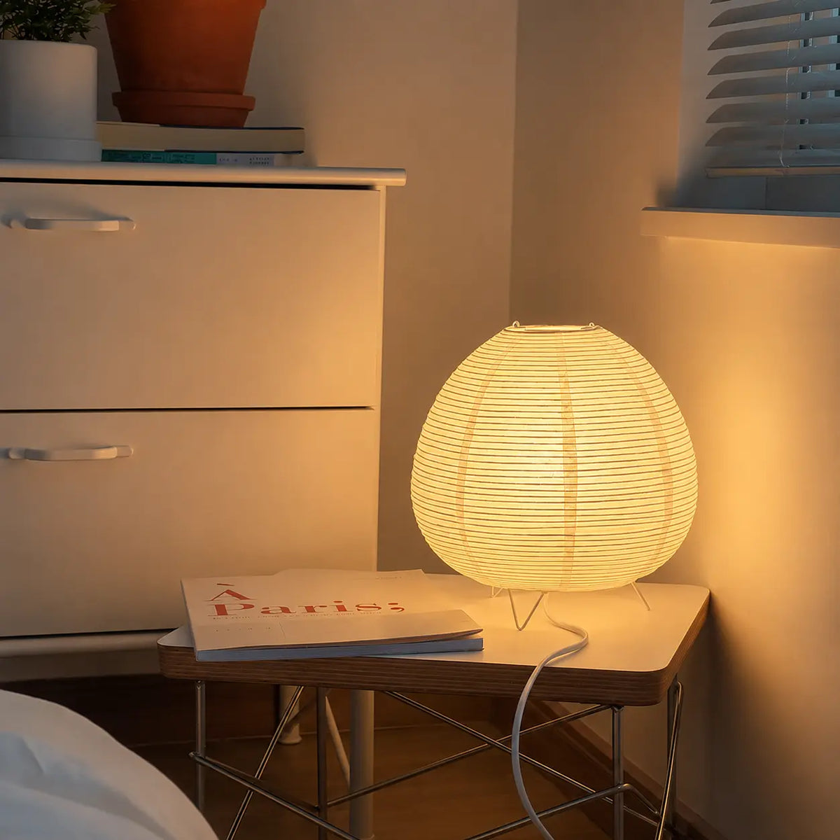 Lampe lanterne japonaise allumée sur une petite table de chevet en bois dans un coin de chambre chaleureux.
