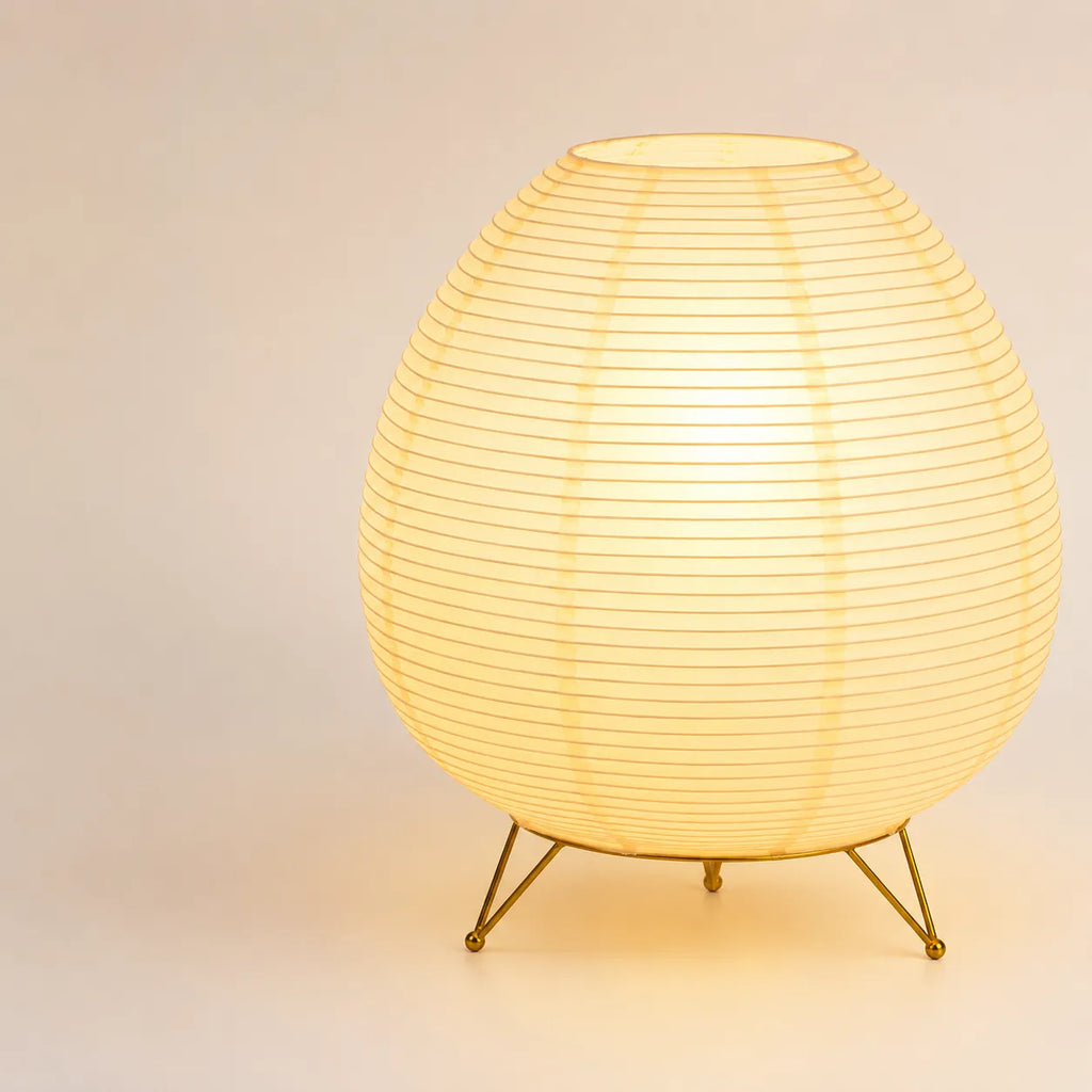 Lampe lanterne japonaise de forme ovale allumée sur son trépied en métal doré brillant.