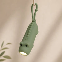 Lampe torche dinosaure verte suspendue par son attache en silicone, éclairant vers le bas.