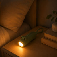 Lampe torche et veilleuse en forme de dinosaure vert posée sur une table de chevet en bois, diffusant une lumière chaleureuse.