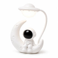 Veilleuse blanche originale mettant en scène un petit astronaute assis sur un croissant de lune texturé. Une tige flexible blanche part de l'arrière pour suspendre une lampe en forme de soucoupe volante illuminée au-dessus du personnage, présentée de trois-quarts face sur fond blanc.