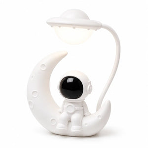 Veilleuse blanche originale mettant en scène un petit astronaute assis sur un croissant de lune texturé. Une tige flexible blanche part de l'arrière pour suspendre une lampe en forme de soucoupe volante illuminée au-dessus du personnage, présentée de trois-quarts face sur fond blanc.