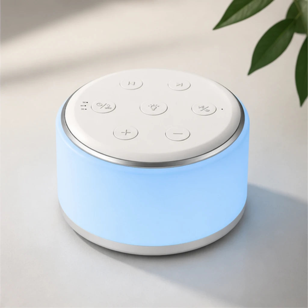 Machine à bruit blanc et veilleuse cylindrique allumée diffusant une lueur bleu clair apaisante. Posée sur une surface neutre à côté de quelques feuilles vertes, sa face supérieure blanche met en avant ses boutons de contrôle épurés.