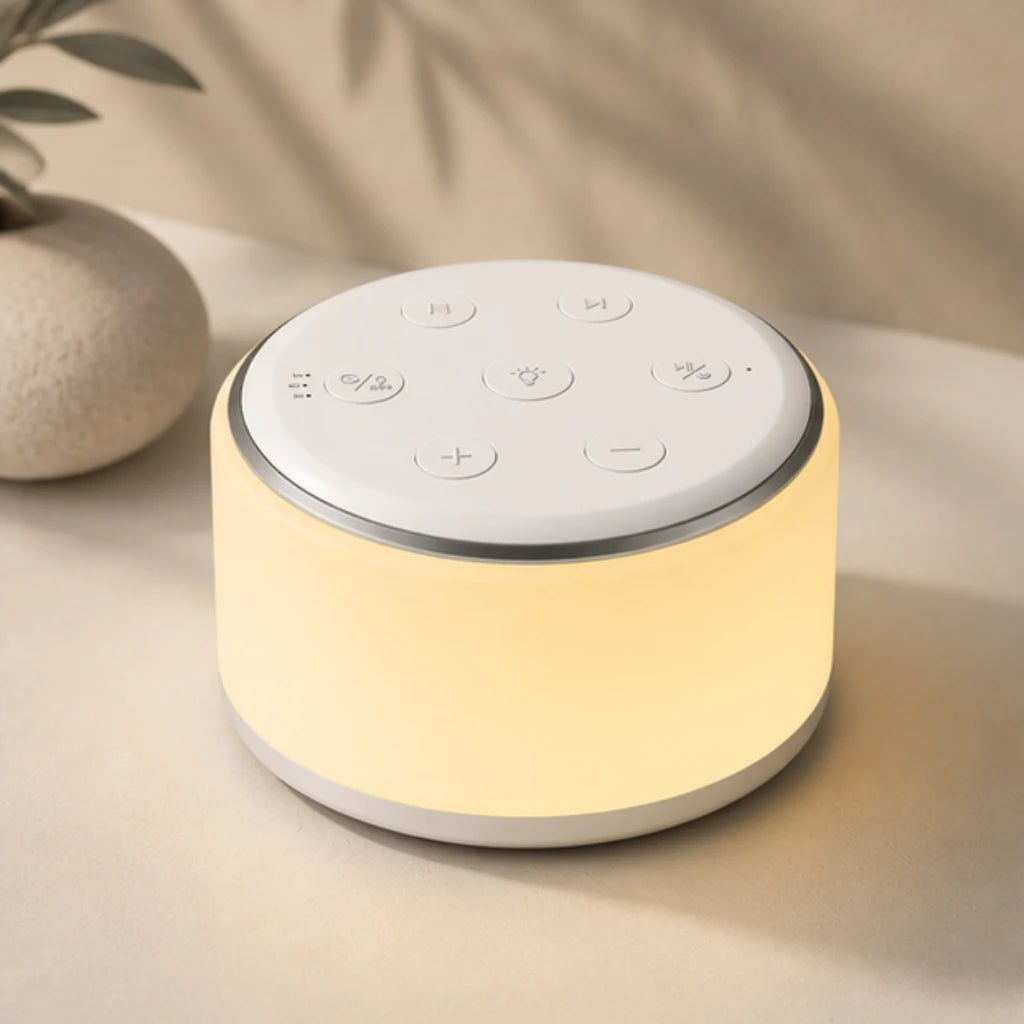 Machine à bruit blanc et veilleuse de forme cylindrique allumée, diffusant une douce lueur jaune tamisée. Elle est présentée en gros plan sur une surface beige épurée à côté d’un petit vase en céramique texturée, baignée dans un jeu d'ombres douces de feuillages en arrière-plan.