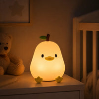 Petite veilleuse poussin éclairée diffusant une lumière chaude sur une table de chevet à côté d'un lit d'enfant et d'un ours en peluche.