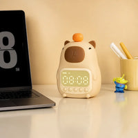 Réveil veilleuse capybara installé sur un bureau d'enfant à côté d'un ordinateur portable et d'un pot à crayons.