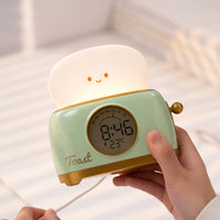 Réveil veilleuse grille-pain vert pastel allumé tenu en main et connecté à son câble de charge USB.
