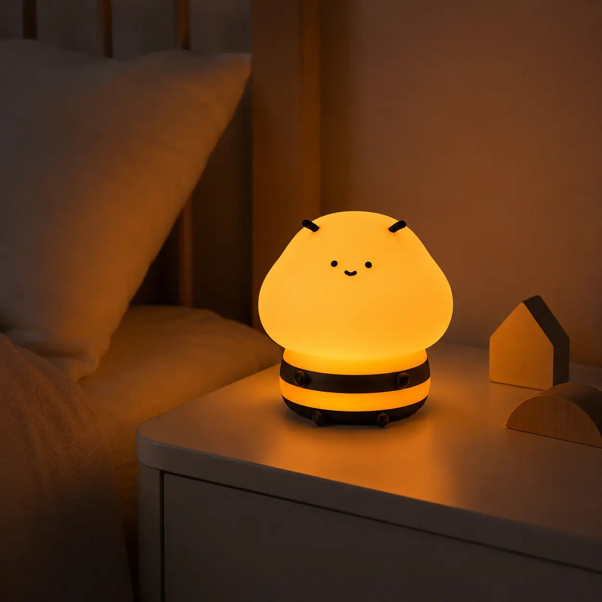 Veilleuse enfant en silicone souple en forme de petite abeille mignonne et souriante, allumée sur une table de chevet blanche dans une chambre, diffusant une douce lueur jaune chaleureuse à côté du lit.