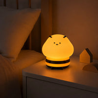 Veilleuse enfant en silicone souple en forme de petite abeille mignonne et souriante, allumée sur une table de chevet blanche dans une chambre, diffusant une douce lueur jaune chaleureuse à côté du lit.