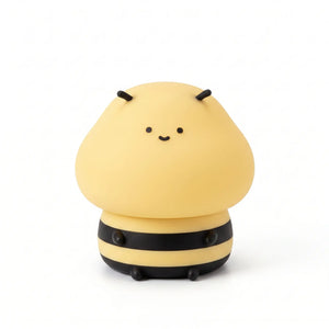 Veilleuse nomade en silicone souple en forme de petite abeille mignonne et souriante, allumée et diffusant une douce lueur jaune avec ses rayures noires caractéristiques, présentée de face sur fond blanc.