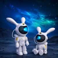 Projecteurs veilleuses astronaute lapin blanc présentés dans les deux versions, debout et assis, sur un sol lunaire avec une galaxie bleue en arrière-plan.