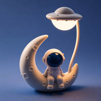 Veilleuse astronaute lune allumée avec un éclairage chaud et doux sur fond bleu nuit.