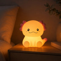 Veilleuse en silicone en forme de petit axolotl mignon allumée, diffusant une lueur chaude et tamisée sur une table de chevet en bois dans une chambre à coucher à l'atmosphère douce et apaisante.