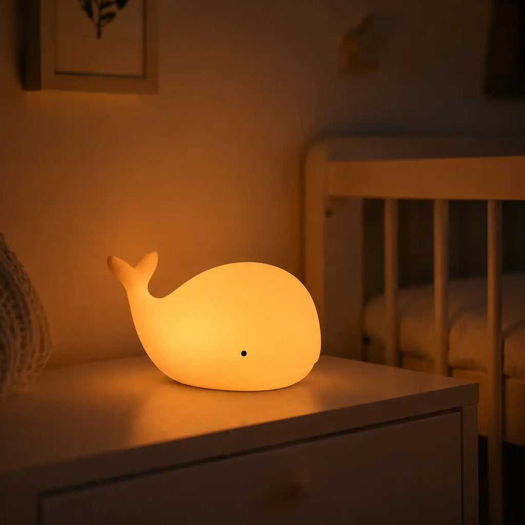 Veilleuse en forme de petite baleine allumée posée sur une table de chevet dans une ambiance chaleureuse.