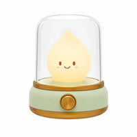Veilleuse bougie LED en forme de petite flamme souriante sous un dôme transparent avec une base verte et dorée.