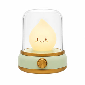 Veilleuse bougie LED en forme de petite flamme souriante sous un dôme transparent avec une base verte et dorée.