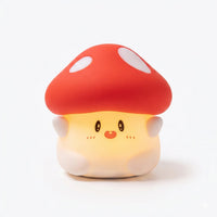 Veilleuse en silicone en forme de petit champignon mignon, allumée sur un fond blanc uni. Le personnage présente un chapeau rouge à grands pois blancs et un pied au visage souriant avec de petits yeux noirs et des joues rosées, diffusant une douce lueur chaude et tamisée.