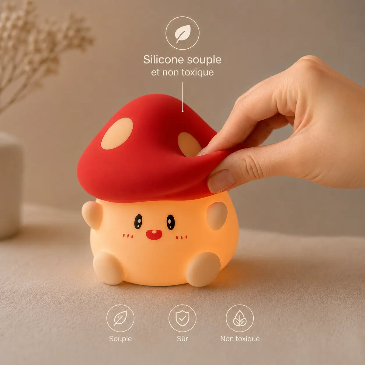 Infographie en français mettant en valeur la texture d'une veilleuse champignon en silicone allumée. Une main attrape et pince délicatement le bord souple de son chapeau rouge pour démontrer sa flexibilité. Un repère textuel indique "Silicone souple et non toxique", complété en bas par trois icônes légendées : Souple, Sûr, et Non toxique.