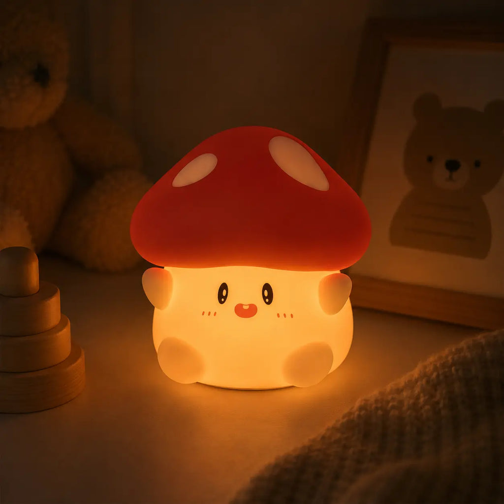 Veilleuse en silicone en forme de petit champignon mignon et allumée, posée dans une chambre d'enfant à l'ambiance tamisée. Elle diffuse une lueur chaude et rassurante sur un meuble, à côté d'un cadre photo en bois illustrant un ours, d'un jeu à empiler en bois et d'une peluche en arrière-plan.