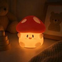 Veilleuse en silicone en forme de petit champignon mignon et allumée, posée dans une chambre d'enfant à l'ambiance tamisée. Elle diffuse une lueur chaude et rassurante sur un meuble, à côté d'un cadre photo en bois illustrant un ours, d'un jeu à empiler en bois et d'une peluche en arrière-plan.