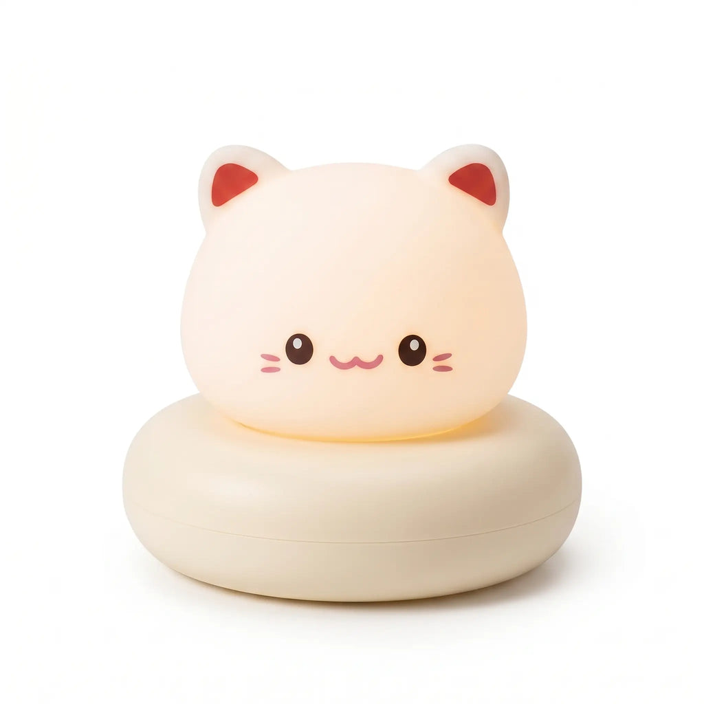 Veilleuse en forme de tête de petit chat blanc allumée, posée sur un large socle circulaire assorti de couleur crème sur fond blanc uni. Le personnage présente un visage blanc lumineux et arrondi avec des petits yeux noirs, un sourire mignon, de petites moustaches et l'intérieur des oreilles teinté de rouge, diffusant une douce lueur chaleureuse.