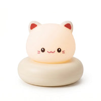 Veilleuse en forme de tête de petit chat blanc allumée, posée sur un large socle circulaire assorti de couleur crème sur fond blanc uni. Le personnage présente un visage blanc lumineux et arrondi avec des petits yeux noirs, un sourire mignon, de petites moustaches et l'intérieur des oreilles teinté de rouge, diffusant une douce lueur chaleureuse.