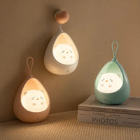 Trois veilleuses chat cocon allumées diffusant une lueur chaleureuse dans une chambre : la blanche est suspendue à une patère en forme de cœur, la rose est posée sur un plaid beige et la bleue repose sur une pile de livres minimalistes.