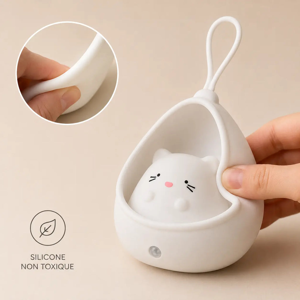Gros plan sur une main pressant le bord souple d'une veilleuse chat cocon blanche éteinte sur fond beige, illustrant sa texture malléable, avec un macaron textuel indiquant silicone non toxique.