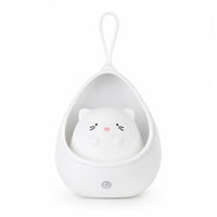 Veilleuse blanche en forme de petit chat mignon installé à l'intérieur d'un cocon protecteur en forme de goutte. La structure intègre une lanière en silicone sur le dessus pour être suspendue facilement et un capteur circulaire sur la base inférieure, présentée de face sur fond blanc.