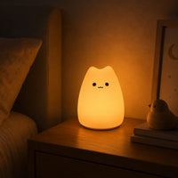 Veilleuse en silicone en forme de petit chat arrondi allumée sur une table de chevet en bois. Elle diffuse une douce lueur chaude orangée à côté d'un lit et de petits objets déco, créant une ambiance calme et propice au sommeil.