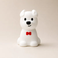 Veilleuse en silicone en forme de petit chien blanc avec un nœud papillon rouge, sur un fond beige neutre.