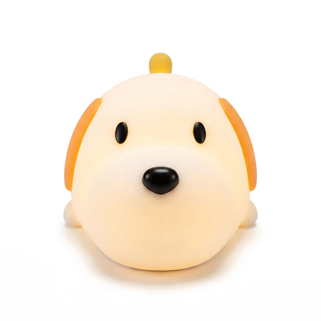 Veilleuse en silicone en forme de tête de petit chien mignon, allumée sur un fond blanc uni. Le personnage présente deux yeux noirs ovales, un nez noir arrondi, des oreilles tombantes orangées et une petite crête ou queue sur le dessus, diffusant une douce lueur chaude et tamisée.