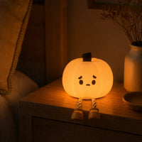 Veilleuse en silicone en forme de petite citrouille mignonne et allumée, assise sur le bord d'une table de chevet en bois à côté d'un lit. Le personnage diffuse une douce lueur chaude et tamisée dans une chambre à coucher à l'ambiance calme, avec ses petites jambes en corde pendant dans le vide, à côté d'un petit vase blanc.