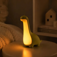 Veilleuse en forme de dinosaure vert allumée posée sur une table de chevet dans une ambiance chaleureuse.