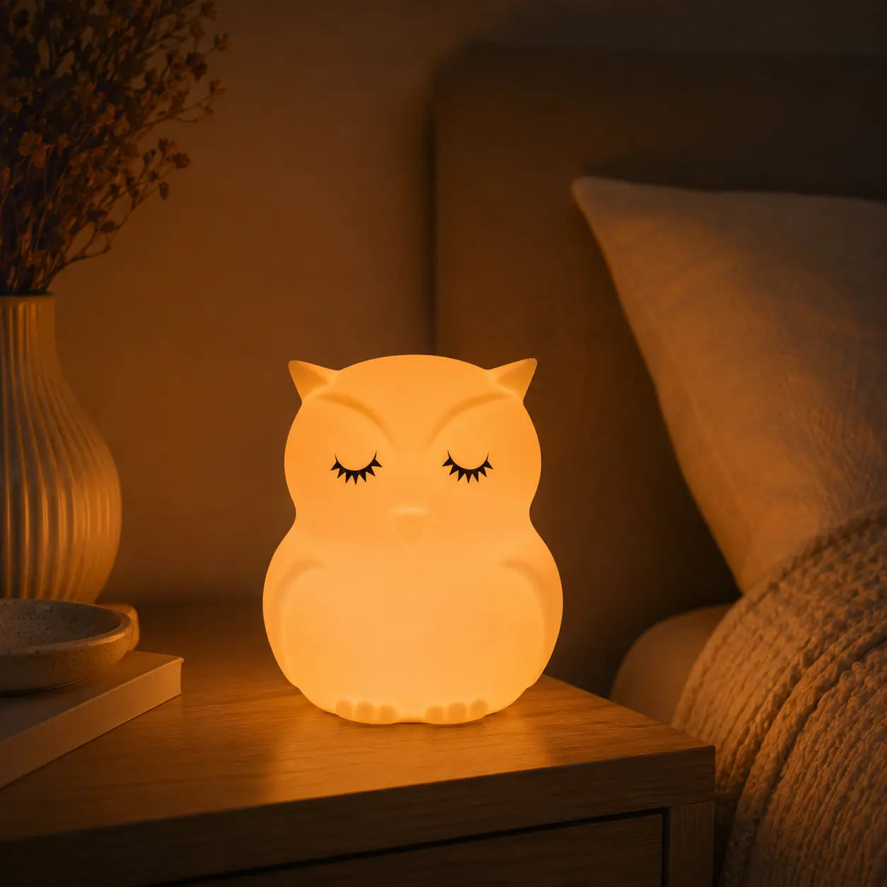 Veilleuse en forme de petit hibou aux yeux fermés allumée posée sur une table de chevet dans une ambiance chaleureuse.