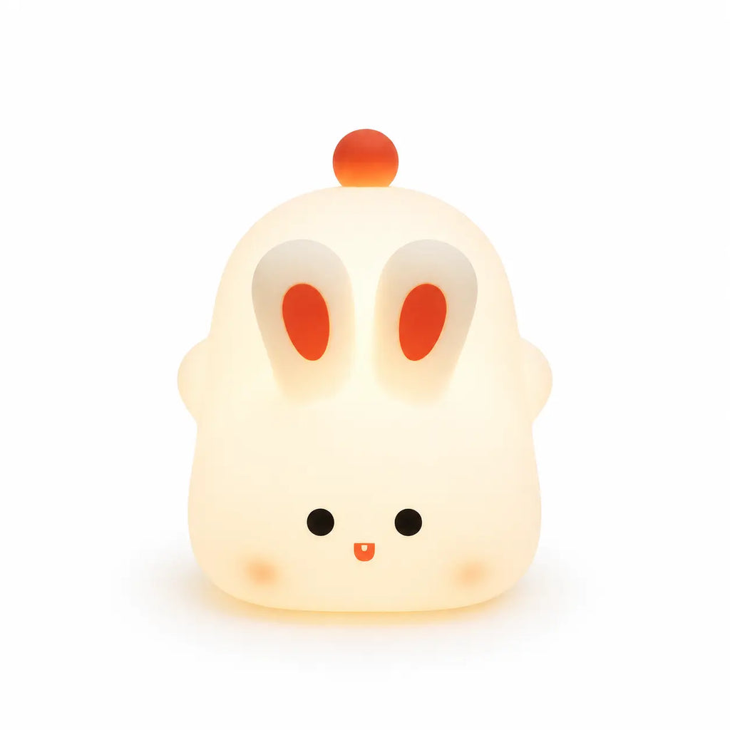 Veilleuse en silicone en forme de petit lapin blanc et arrondi allumée, diffusant une douce lueur jaune tamisée. Elle se distingue par ses deux grandes oreilles dressées aux centres orange, ses petits yeux ronds, sa bouche souriante, ses joues délicatement rosées et un petit pompon rouge sur le dessus de la tête, présentée de face sur fond blanc.