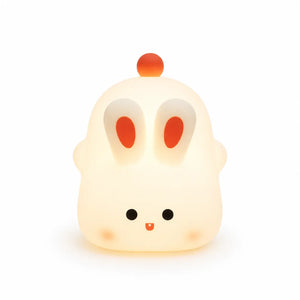 Veilleuse en silicone en forme de petit lapin blanc et arrondi allumée, diffusant une douce lueur jaune tamisée. Elle se distingue par ses deux grandes oreilles dressées aux centres orange, ses petits yeux ronds, sa bouche souriante, ses joues délicatement rosées et un petit pompon rouge sur le dessus de la tête, présentée de face sur fond blanc.