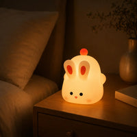 Veilleuse en silicone en forme de petit lapin blanc allumée sur une table de chevet en bois dans une chambre, diffusant une douce lueur jaune tamisée. Le lapin, doté de deux grandes oreilles aux centres orange et d'un petit pompon rouge sur la tête, est posé à côté d'un lit et d'un vase pour créer une ambiance chaleureuse et apaisante.