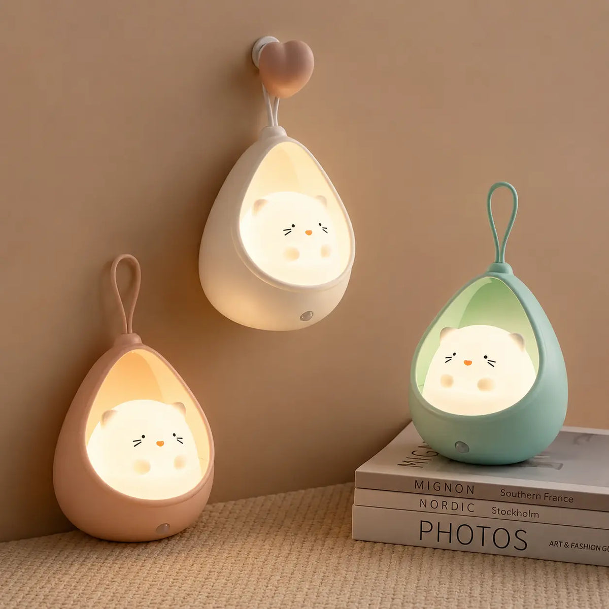 Trois veilleuses lapin cocon allumées diffusant une lueur chaleureuse : le modèle blanc est suspendu à une patère en forme de cœur, le rose est posé au sol sur un plaid beige, et le bleu repose sur une pile de livres minimalistes.