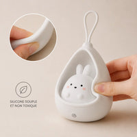 Gros plan sur une main pressant le silicone blanc et souple d'une veilleuse lapin cocon éteinte sur fond beige, illustrant sa texture malléable avec une icône de feuille et l'inscription "silicone souple et non toxique".