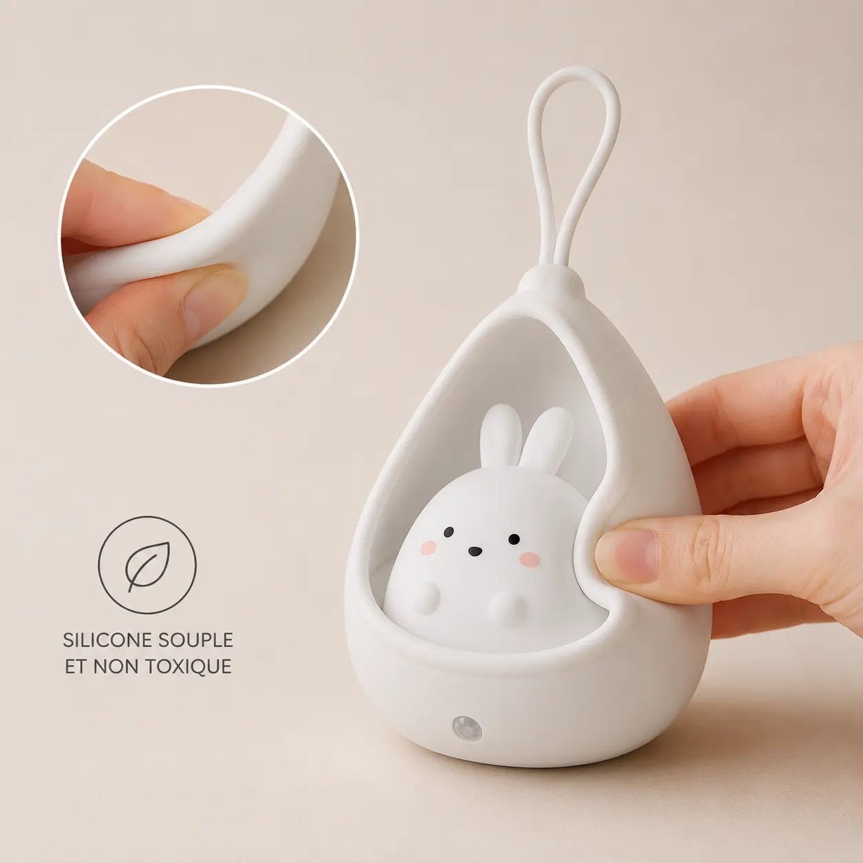 Gros plan sur une main pressant le silicone blanc et souple d'une veilleuse lapin cocon éteinte sur fond beige, illustrant sa texture malléable avec une icône de feuille et l'inscription "silicone souple et non toxique".