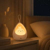 Veilleuse lapin cocon blanche allumée sur une table de chevet en bois dans une chambre de bébé ou d'adulte. Elle diffuse une lumière chaude tamisée à côté d'un lit douillet et d'un vase blanc avec des fleurs séchées, sous une fenêtre ouverte sur une nuit étoilée.