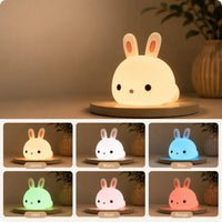 Composition d’images présentant une veilleuse lapin en silicone allumée sur un socle rond, diffusant une lueur jaune chaleureuse. Au bas, six vignettes étiquetées illustrent les différentes variations de couleurs de sa lumière : Jaune, Blanc, Bleu, Vert, Rose et Rouge, dans une ambiance intérieure minimaliste.